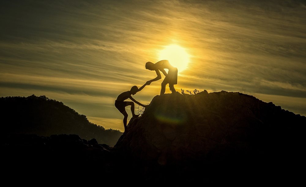 Silhouettes of one man helping a young boy climb a hill.