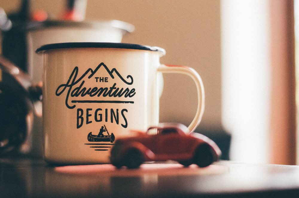 A mug reading "the adventure begins", with a toy car besides it.