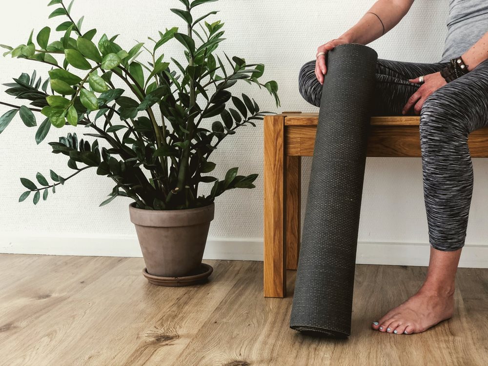 Person sitting next to a plant, holding a yoga mat.